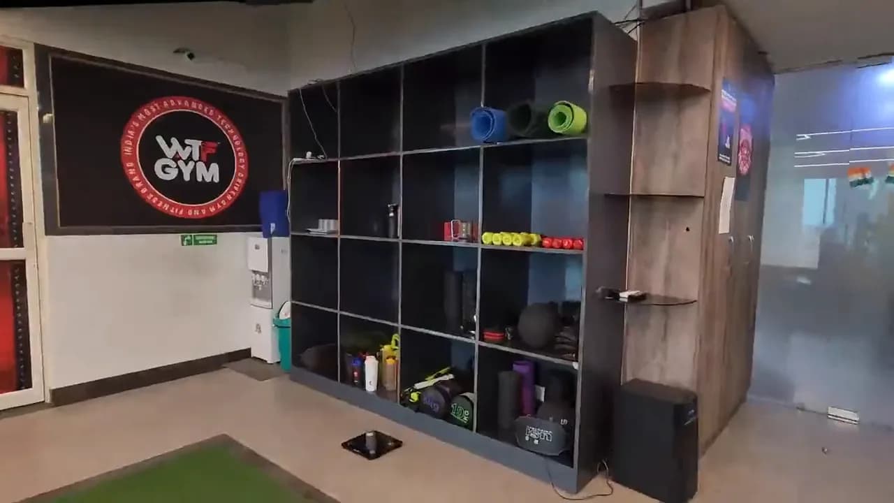 A gym corner: an accessory rack of mats, rollers and dumbbells beside a red-and-black wall emblem, turf at its foot